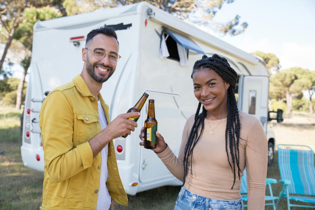 Smiling couple holds beer bottles in front of an RV, enjoying a sunny day outdoors.